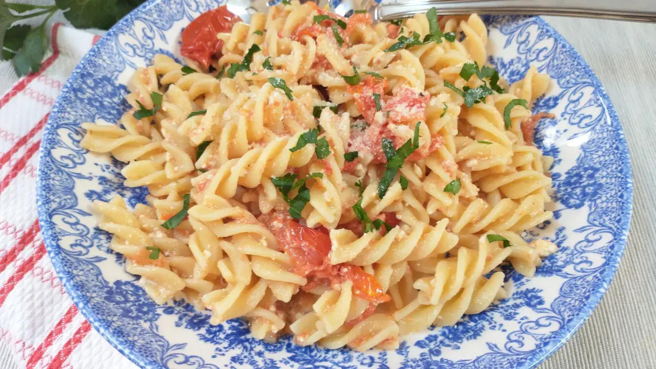 Fusilli z pomidorkami koktajlowymi i serem feta, posypane natką pietruszki, na talerzu z niebieskim wzorem.