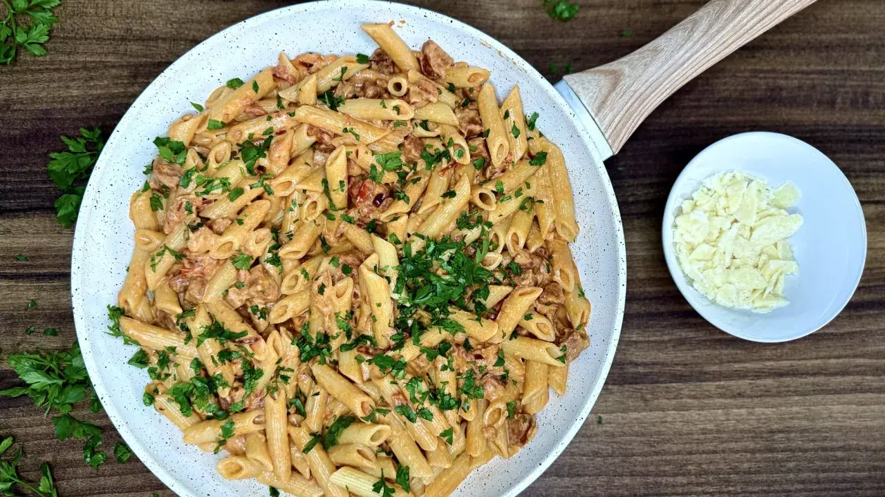 Penne z kremowym sosem i kawałkami pikantnego chorizo, posypane świeżą natką pietruszki. Obok miseczka z parmezanem.