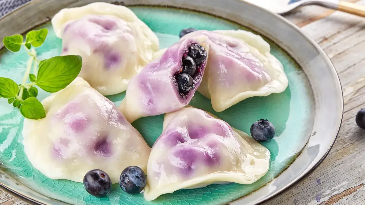 Pyszne pierogi z jagodami na turkusowym talerzu, ozdobione świeżymi borówkami i gałązką ziół.