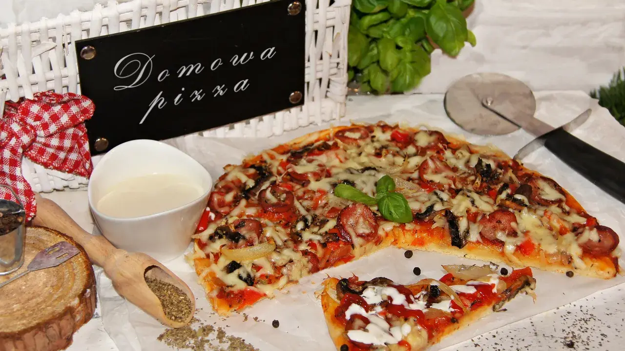 Domowa pizza bez drożdży, z kiełbasą i warzywami, prezentowana na białym papierze, obok sosu i przypraw.