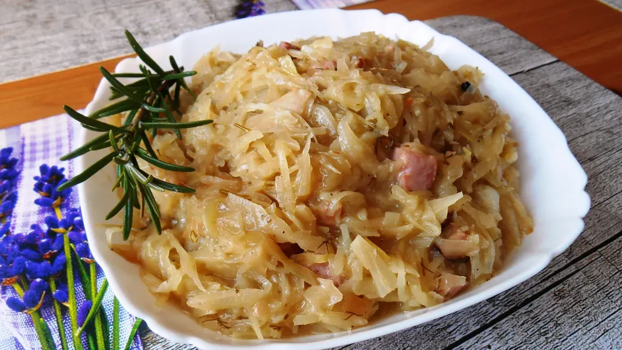 Młoda kapusta zasmażana z boczkiem, podana na białym talerzu, ozdobiona gałązką rozmarynu.