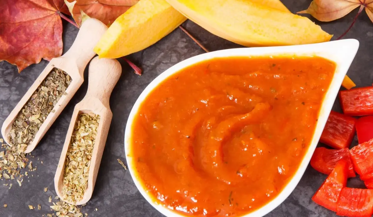 Pomarańczowy, gęsty ketchup z dyni w białej miseczce, obok drewniane łyżeczki z ziołami, kawałki papryki i jesienne liście.