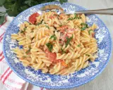 Fusilli z pomidorkami koktajlowymi i serem feta, posypane natką pietruszki, na talerzu z niebieskim wzorem.