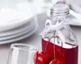 Elegancka nalewka z wiśni w butelce, ozdobiona wstążką i świeżymi owocami. Obok stoi kieliszek, a w tle stos talerzy.