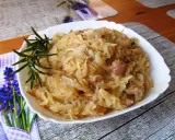 Młoda kapusta zasmażana z boczkiem, podana na białym talerzu, ozdobiona gałązką rozmarynu.