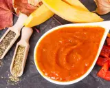 Pomarańczowy, gęsty ketchup z dyni w białej miseczce, obok drewniane łyżeczki z ziołami, kawałki papryki i jesienne liście.