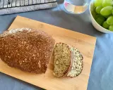 Chleb z twarogu, pełen ziaren i orzechów, pokrojony na desce. Obok miska zielonych winogron i szklanka wody.