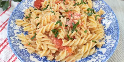 Fusilli z pomidorkami koktajlowymi i serem feta, posypane natką pietruszki, na talerzu z niebieskim wzorem.