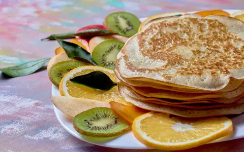 Stos puszystych naleśników z budyniem, udekorowany plasterkami kiwi, pomarańczy i jabłka.