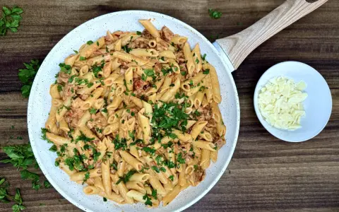Penne z kremowym sosem i kawałkami pikantnego chorizo, posypane świeżą natką pietruszki. Obok miseczka z parmezanem.