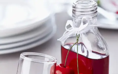 Elegancka nalewka z wiśni w butelce, ozdobiona wstążką i świeżymi owocami. Obok stoi kieliszek, a w tle stos talerzy.