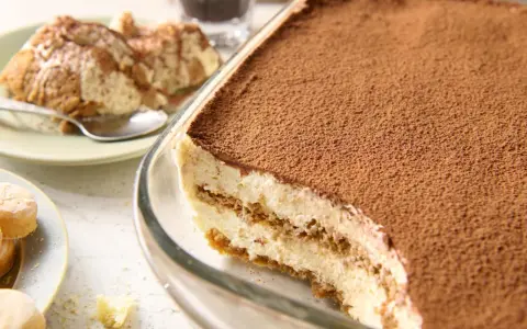Pyszne tiramisu bez jajek w szklanej formie, posypane kakao. Obok porcja deseru na talerzyku.