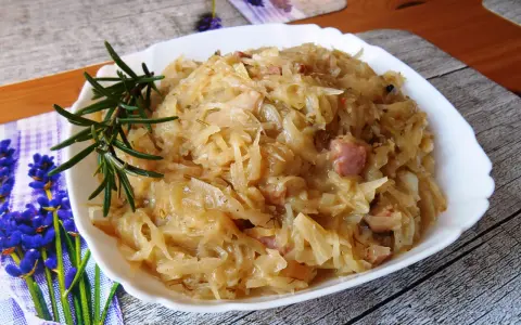 Młoda kapusta zasmażana z boczkiem, podana na białym talerzu, ozdobiona gałązką rozmarynu.