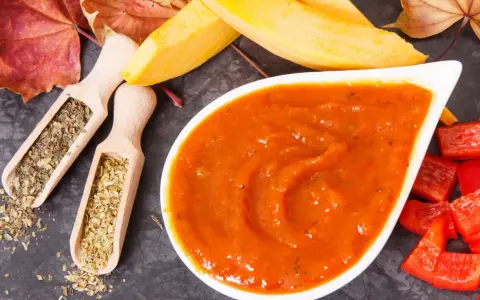 Pomarańczowy, gęsty ketchup z dyni w białej miseczce, obok drewniane łyżeczki z ziołami, kawałki papryki i jesienne liście.