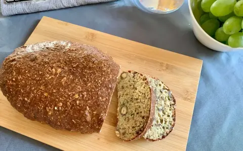 Chleb z twarogu, pełen ziaren i orzechów, pokrojony na desce. Obok miska zielonych winogron i szklanka wody.