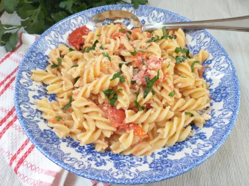 Fusilli z pomidorkami koktajlowymi i serem feta, posypane natką pietruszki, na talerzu z niebieskim wzorem.