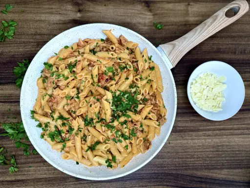 Penne z kremowym sosem i kawałkami pikantnego chorizo, posypane świeżą natką pietruszki. Obok miseczka z parmezanem.