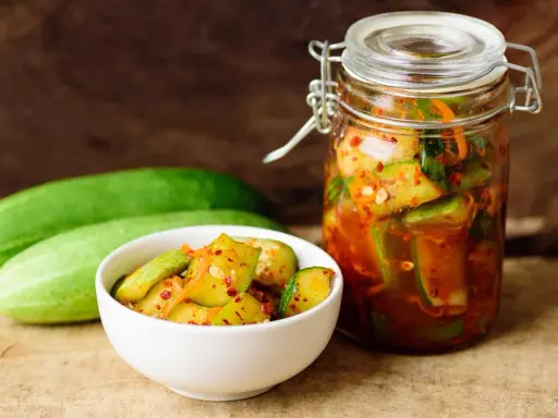 Pikantne ogórki z papryką chili w słoiku i miseczce, obok świeże ogórki.