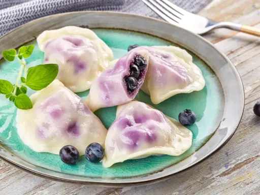 Pyszne pierogi z jagodami na turkusowym talerzu, ozdobione świeżymi borówkami i gałązką ziół.