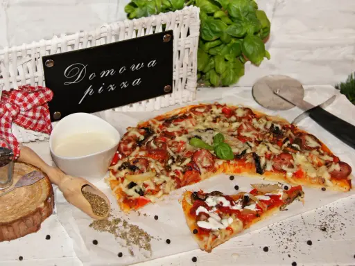 Domowa pizza bez drożdży, z kiełbasą i warzywami, prezentowana na białym papierze, obok sosu i przypraw.