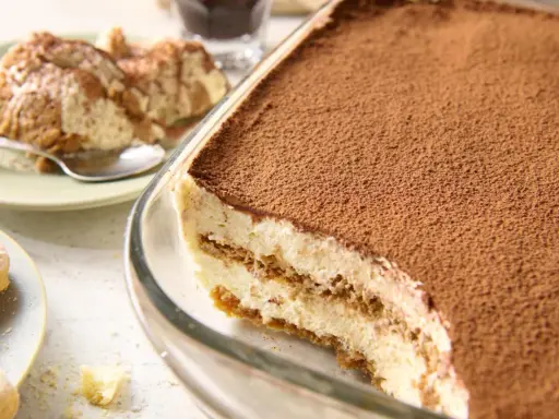 Pyszne tiramisu bez jajek w szklanej formie, posypane kakao. Obok porcja deseru na talerzyku.