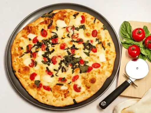 Pyszna pizza z serka wiejskiego, pomidorkami koktajlowymi i bazylią. Obok leżą świeże pomidory i krajalnica do pizzy.