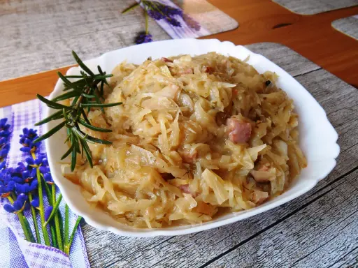 Młoda kapusta zasmażana z boczkiem, podana na białym talerzu, ozdobiona gałązką rozmarynu.