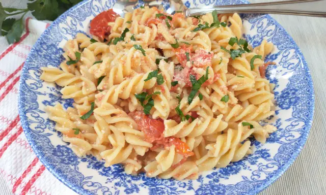 Fusilli z pomidorkami koktajlowymi i serem feta, posypane natką pietruszki, na talerzu z niebieskim wzorem.