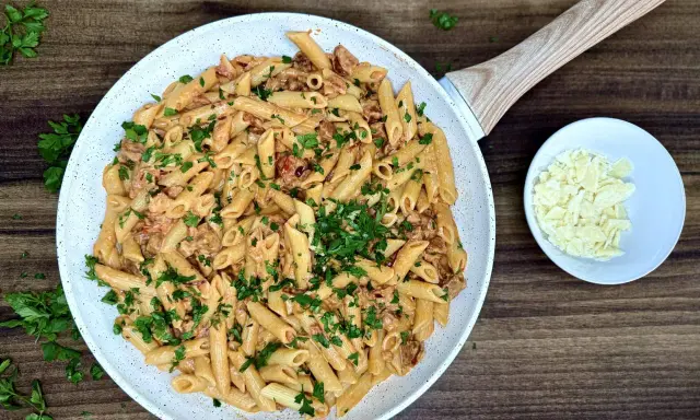 Penne z kremowym sosem i kawałkami pikantnego chorizo, posypane świeżą natką pietruszki. Obok miseczka z parmezanem.