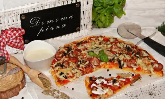 Domowa pizza bez drożdży, z kiełbasą i warzywami, prezentowana na białym papierze, obok sosu i przypraw.