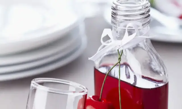 Elegancka nalewka z wiśni w butelce, ozdobiona wstążką i świeżymi owocami. Obok stoi kieliszek, a w tle stos talerzy.
