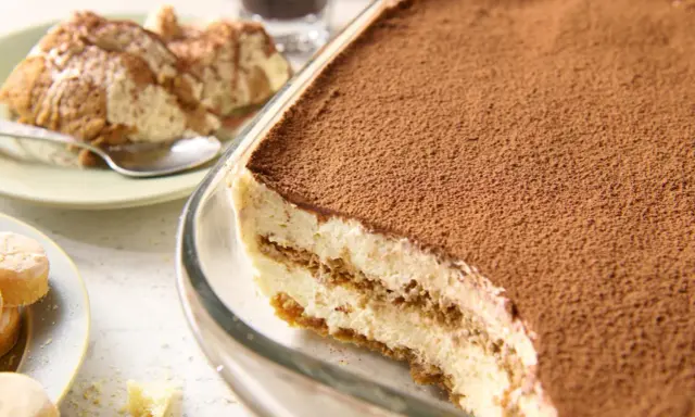 Pyszne tiramisu bez jajek w szklanej formie, posypane kakao. Obok porcja deseru na talerzyku.