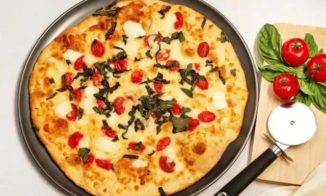 Pyszna pizza z serka wiejskiego, pomidorkami koktajlowymi i bazylią. Obok leżą świeże pomidory i krajalnica do pizzy.