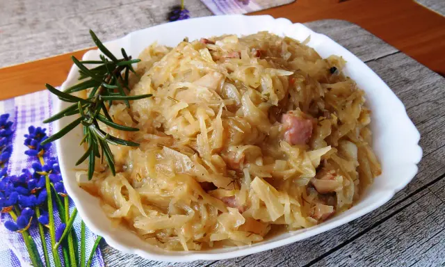 Młoda kapusta zasmażana z boczkiem, podana na białym talerzu, ozdobiona gałązką rozmarynu.