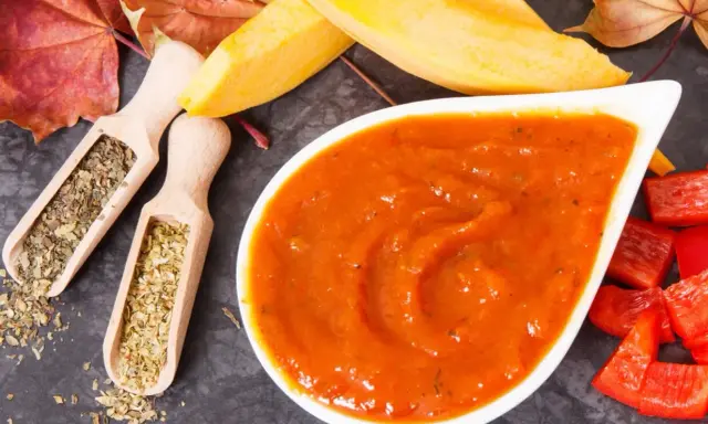Pomarańczowy, gęsty ketchup z dyni w białej miseczce, obok drewniane łyżeczki z ziołami, kawałki papryki i jesienne liście.