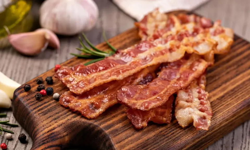 Chrupiący boczek na desce, idealny jako marynata do boczku na grilla. Obok czosnek, rozmaryn i kolorowy pieprz.