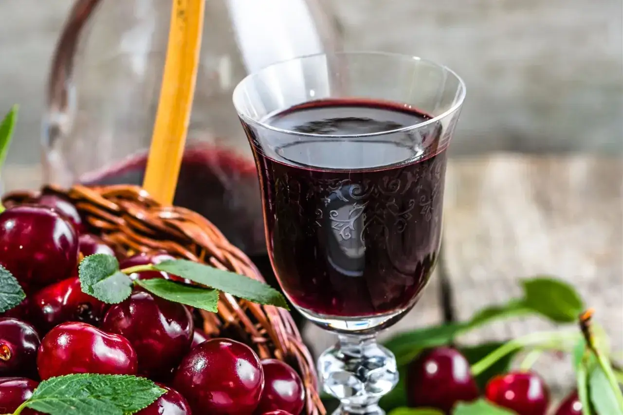 Sok z wiśni przepis babci. Kieliszek ciemnego, aromatycznego soku z wiśni obok koszyka pełnego świeżych owoc&oacute;w.