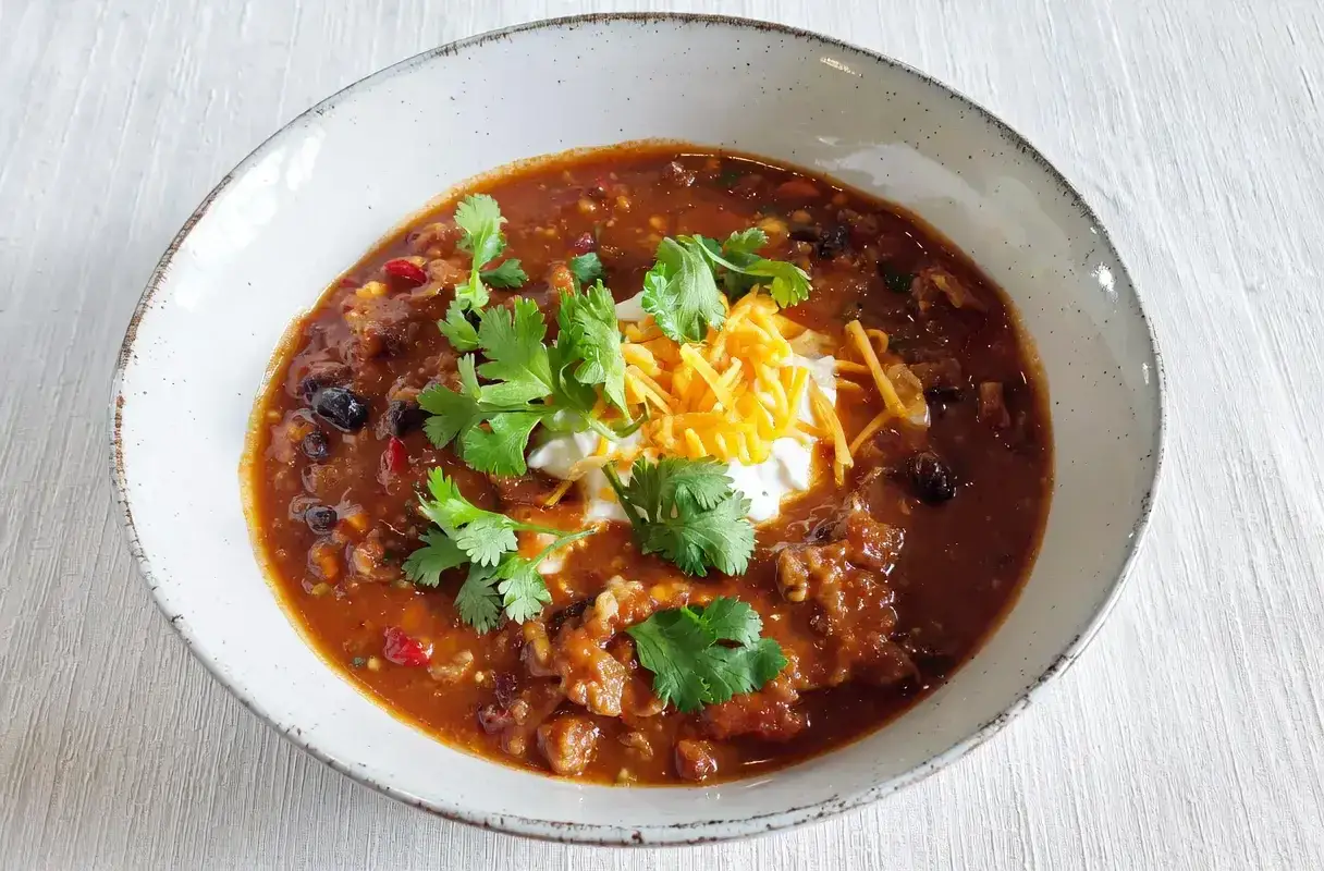 Aromaticzne chili con carne z fasolą, mięsem i przyprawami, udekorowane śmietaną, startym serem i świeżą kolendrą.