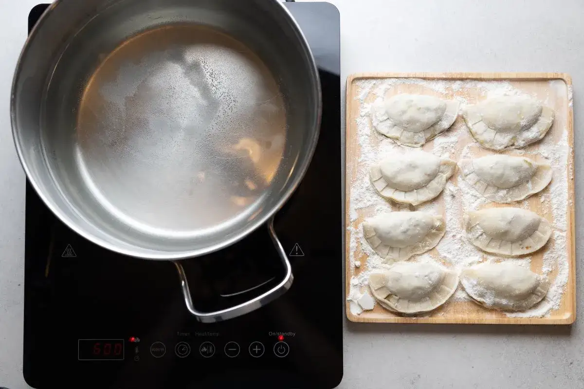 Gotowe do gotowania pierogi z kapustą i grzybami leżą na desce, obok garnka z bulgoczącą wodą.