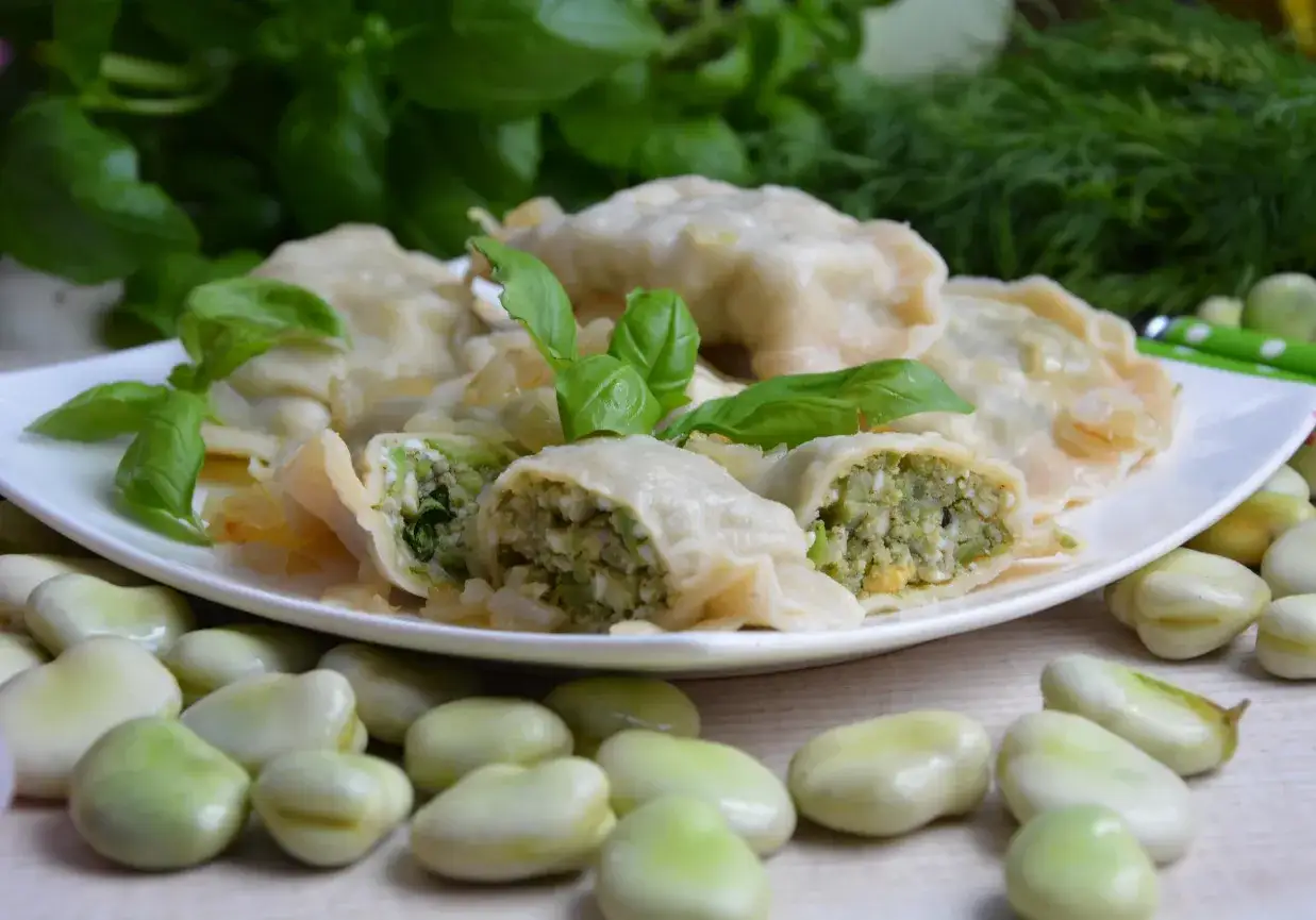 Pyszne pierogi z bobem, jajkiem i ziołami, udekorowane listkami bazylii. Na talerzu widać też świeży bób.