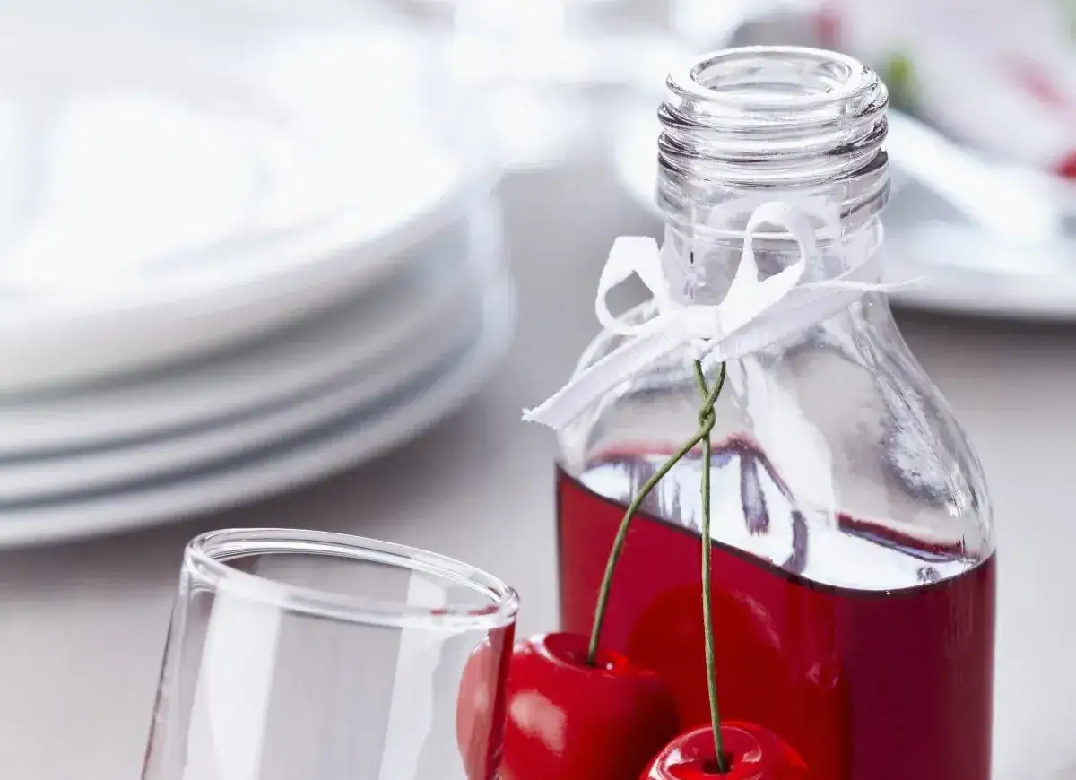 Elegancka nalewka z wiśni w butelce, ozdobiona wstążką i świeżymi wiśniami. Obok stoi pusty kieliszek, a w tle piętrzą się białe talerze.