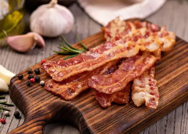 Chrupiący boczek na desce, idealny jako marynata do boczku na grilla. Obok czosnek, rozmaryn i kolorowy pieprz.